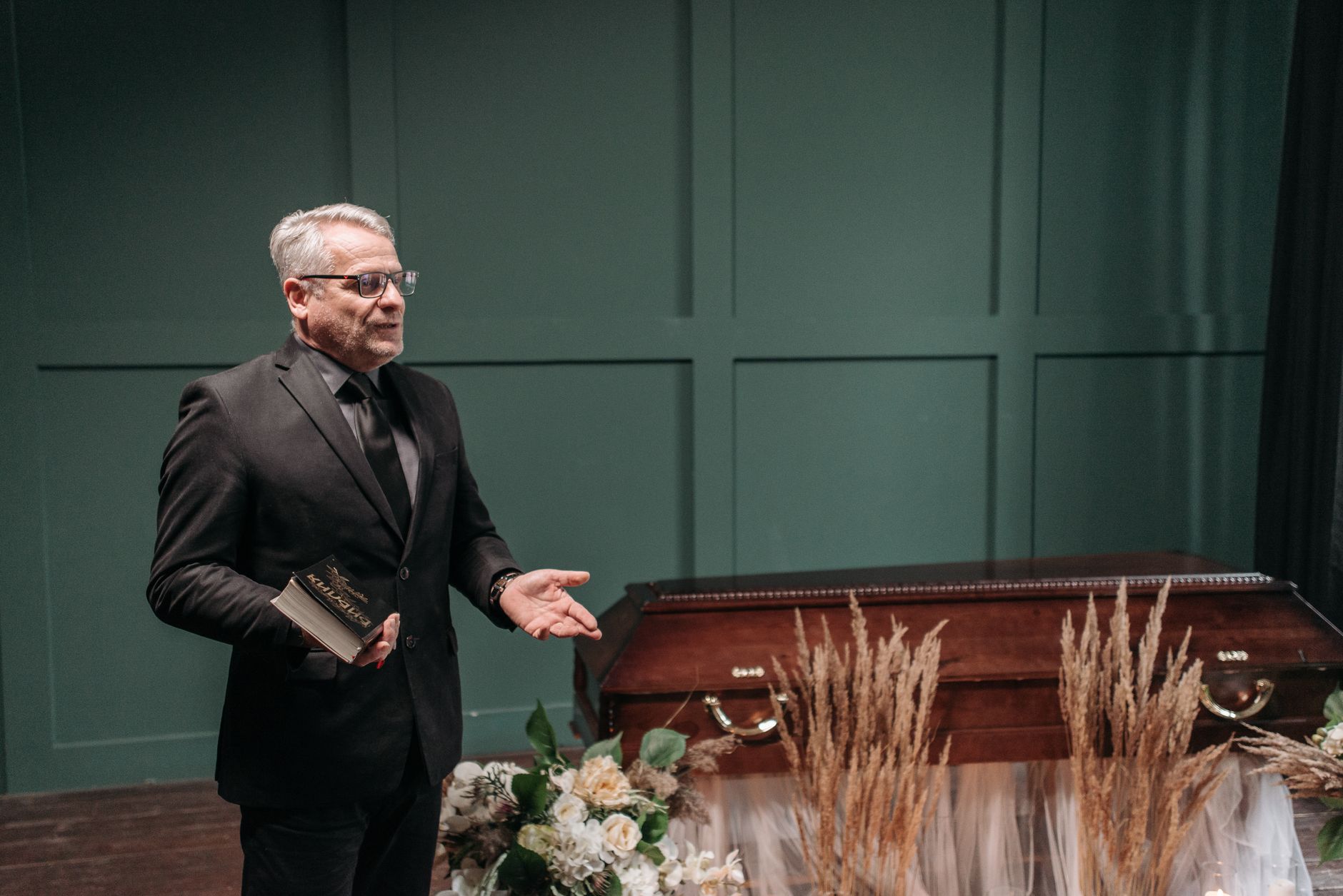 A funeral director speaking at a service with a casket and floral arrangements